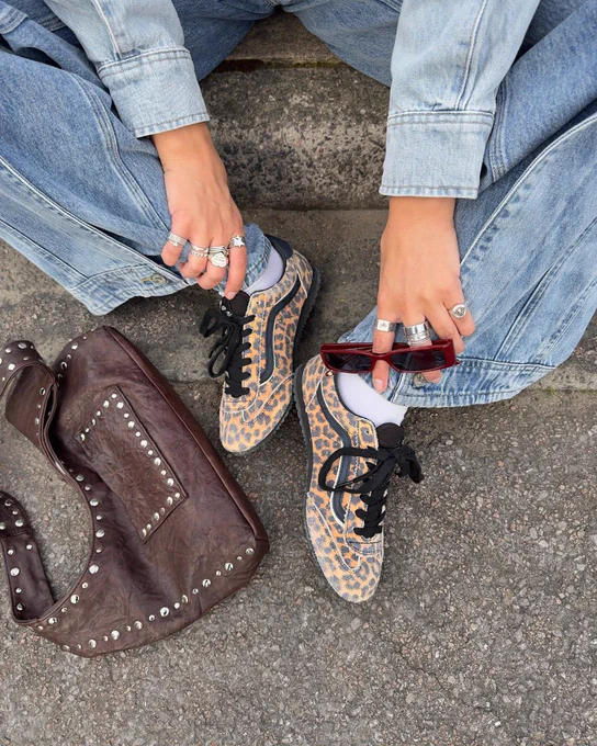 An image of a person's lower legs wearing the same shoes whilst sitting on a pavement next to a brown handbag.