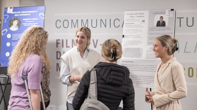 A group of four people stand in discussion near a display area. One person is near a poster titled "Tracking Your Ethical Values" by Stephen Brad. Another nearby display features information about transparency and integrity.