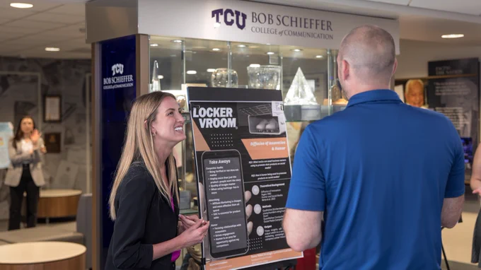 This image shows two people interacting near a display titled "Locker Vroom" in the TCU Bob Schieffer College of Communication. The display details a project or presentation. Another person is visible in the background, standing near glass trophies.