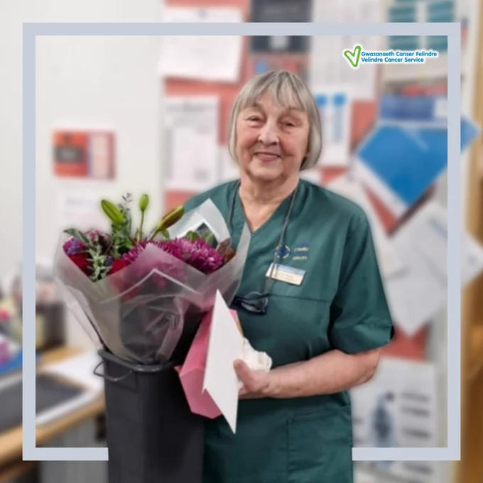 A healthcare worker in green scrubs holding a bouquet of flowers and a card.