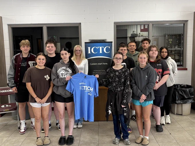 A group of students in casual wear posing together; a girl holds a blue t-shirt with text.