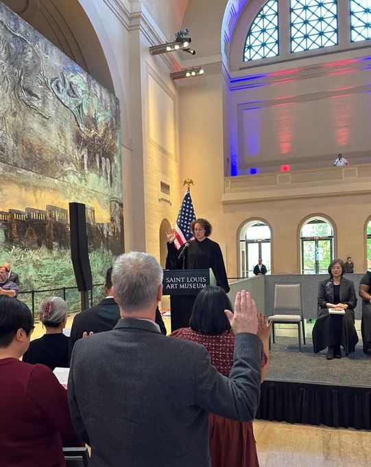 Judge Lanahan in her black robe stands at the podium raising her right hand, the applicants for citizenship also raise their right hands as they take the oath to be come U.S. citizens. In the background there is a large painting that is part of an exhibit at the Saint Louis Art Museum.