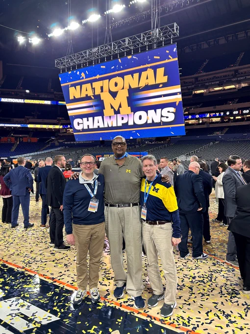 Brian Boesch, left, stands with the broadcast crew after Michigan Men's Basketball won the National Championship, with a large screen in the back reading "National Champions" with blue and yellow confetti covering the court.
