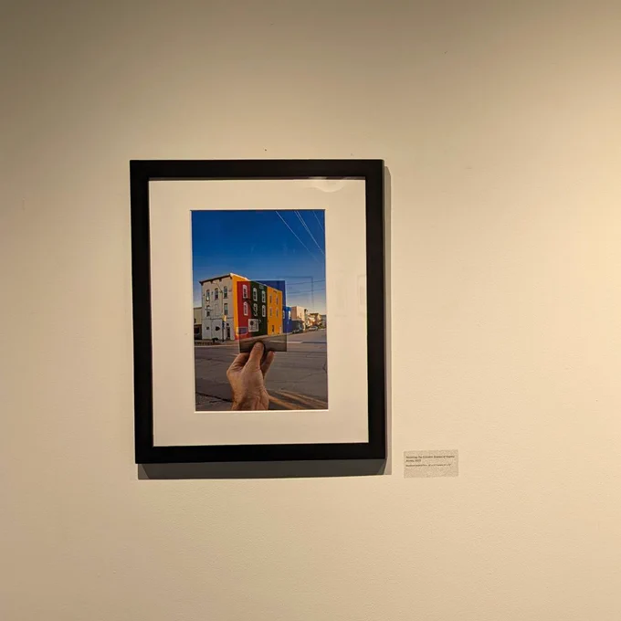 A photograph hanging on a white gallery wall. The photograph features a street with a building, and a hand holding a secondary photo. The secondary photo features a brightly colored building.