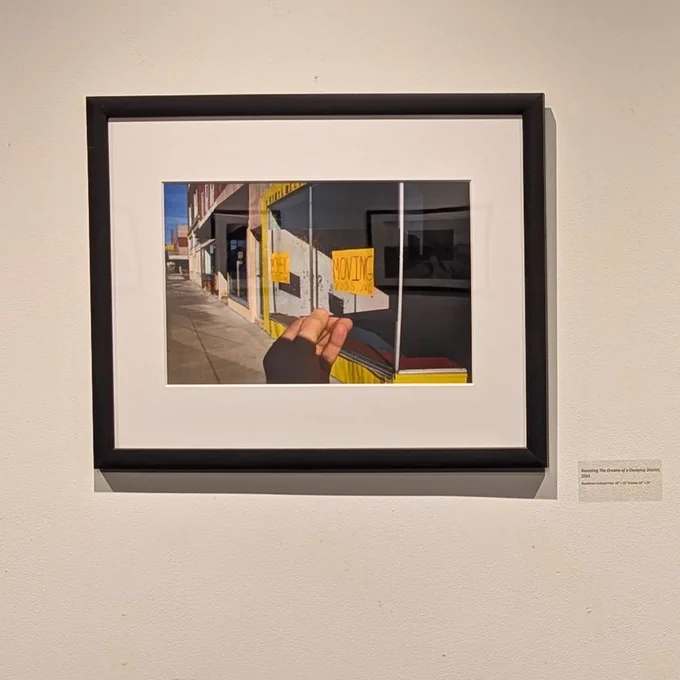 A framed photograph hanging on a plain white wall. The photograph features a hand holding another photograph. The background photo is of a street scene with a sidewalk and several storefronts.