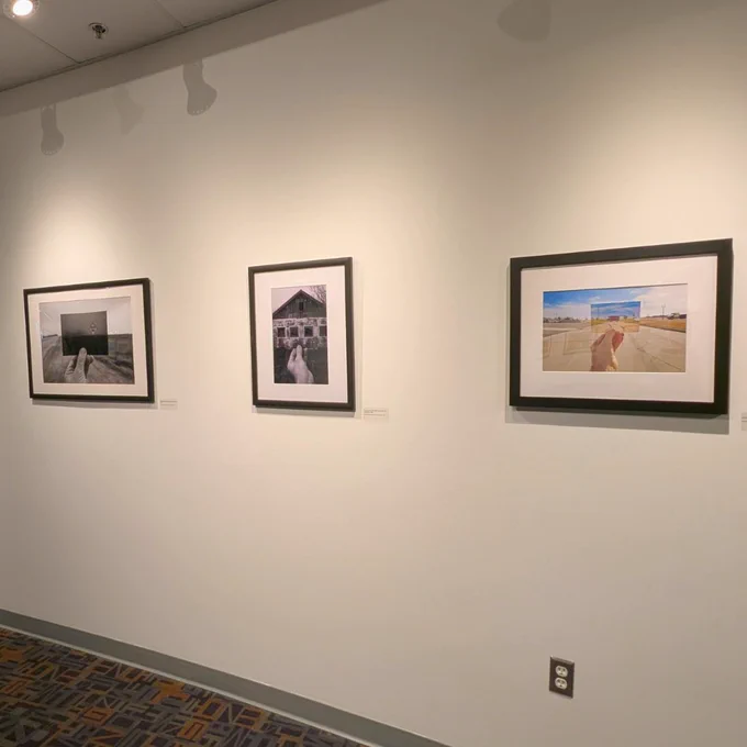A section of a gallery wall displaying three framed photographs. The wall is painted white, and the photographs are arranged horizontally. The first photo on the left is in black and white, depicting an outdoor scene with a hand holding a secondary photo. The central photograph is also black and white, showing a hand holding a secondary photograph, the subject is a rustic looking building. The photograph on the right is in color and displays a hand holding a secondary photograph, there is a wide open road with blue skies.