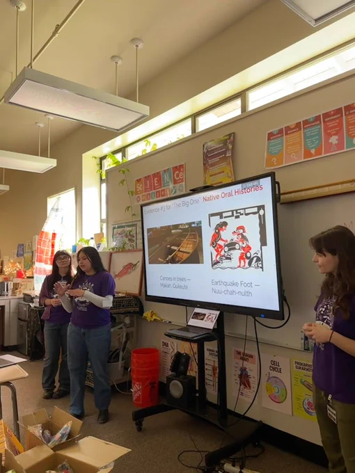 Three people stand at the front of a highschool science classroom, next to a TV. One person speaks towards the class (out of view), while the TV describes "Evidence for 'The Big One': Native Oral Histories" and lists stories about canoes in trees and the earthquake foot.