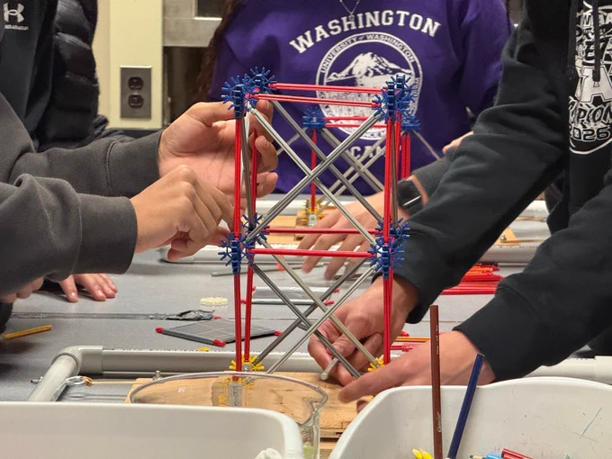 A close up shot of a few students' hands while they work on building structures out of plastic toy building materials. The structures sit on top of a wooden board that is designed to imitate an earthquake when manually shook.
