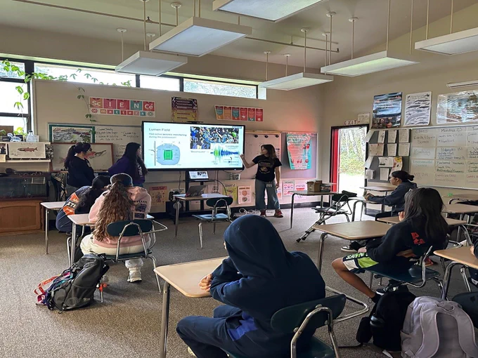 A high school science classroom, with a few rows of students sitting in desks facing the front. At the front, a member of the CCASE team points towards a TV showing seismic activity at Lumen Field during a Seahawks game. Two other team members stand nearby watching.