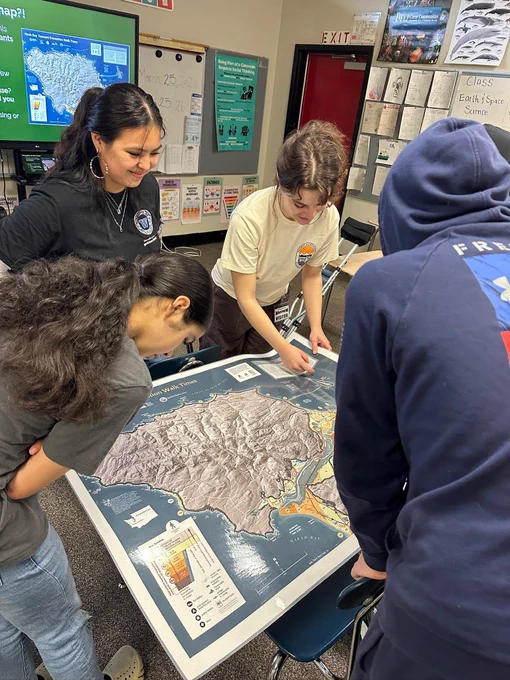 Four people stand around a large map laid out on a school desk in a high school classroom. It is a topographical map of Neah Bay, with tsunami evacuation routes, evacuation time estimates, and tsunami inundation zones outlined.