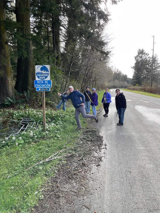 Six people stand at the side of a forested road smiling and holding garden tools, next to a tsunami evacuation route sign that points towards a trail going into the forest. One man in the foreground stands in a running pose, pretending to run up the trail.