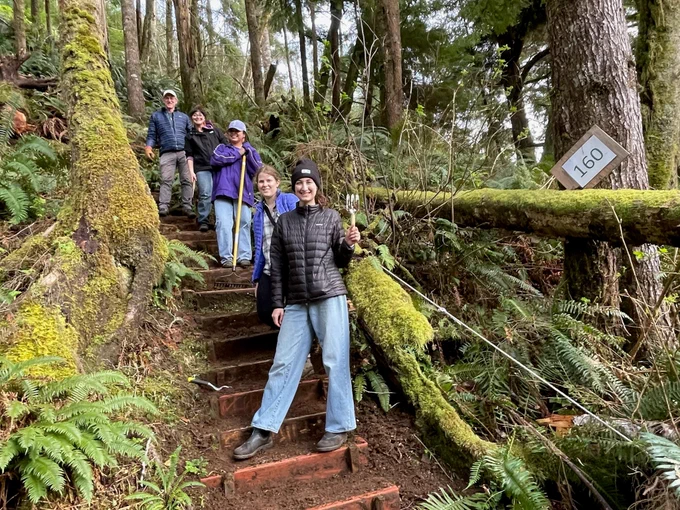 Five people stand in a line on a trail leading through a forest. They are standing on a steep wooden staircase while holding garden tools. A sign denoting 160 feet in elevation hangs from a tree next to the trail.