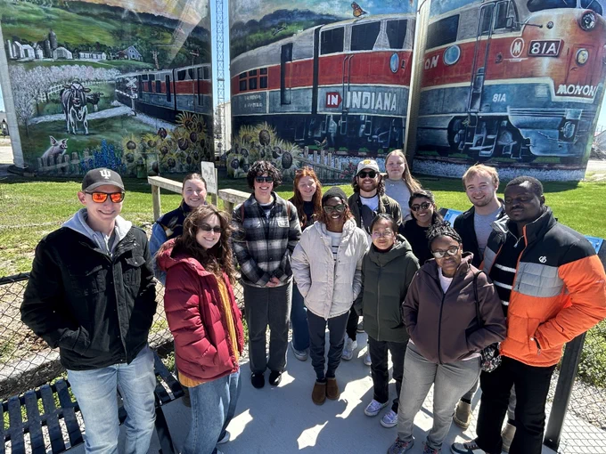 A group of students stand outdoors in front of a colorful mural depicting trains and scenic landscapes. The mural includes the text "Indiana" and "Monon". 