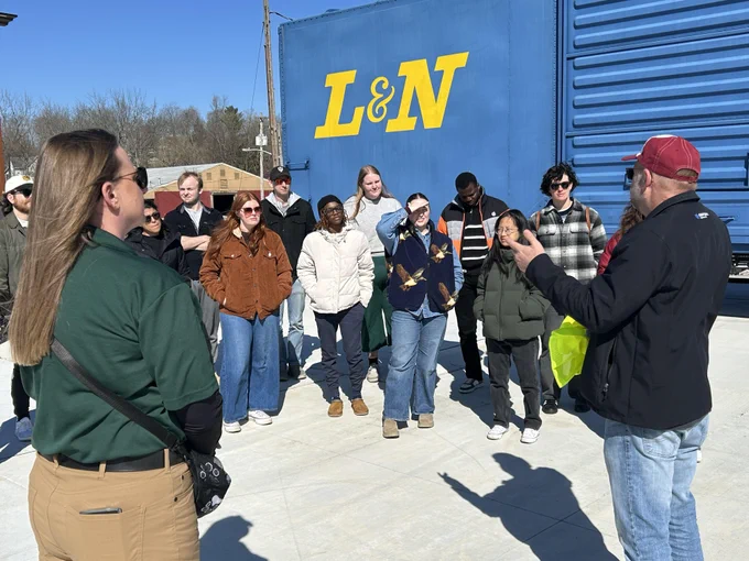 A group of people stands outdoors in a sunny area during the day, listening to a person speaking. Behind them is a large blue structure with "L & N" written in yellow letters.