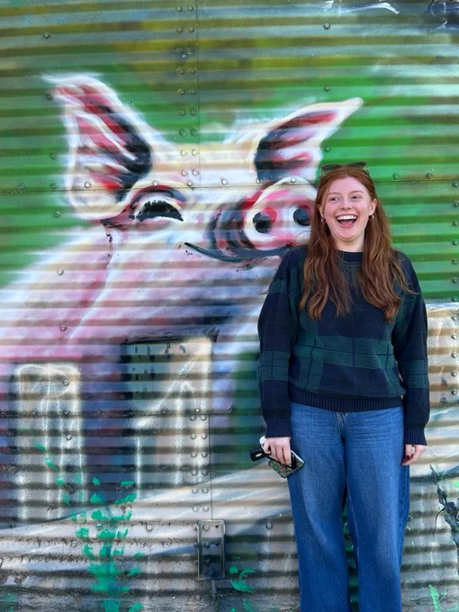 A student stands smiling in front of a vibrant mural featuring a large, detailed painting of a pig with a colorful background.