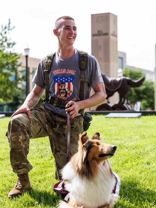 A cadet in camouflage pants kneels on grass, Reveille's leash. The cadet wears a Texas A&M t-shirt, and a Texas A&M University monument is visible in the background.