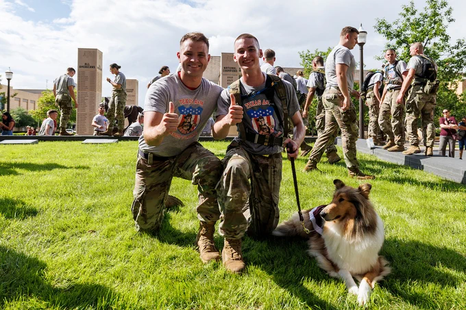 Two Texas A&M cadets in military uniforms kneel on grass next to Reveille. They are giving thumbs up. In the background, more people in similar attire are gathered near memorials. It is a sunny day.