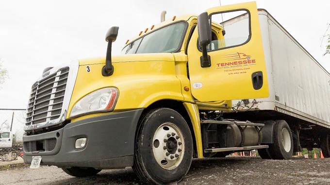 Yellow semi-truck with an open driver's door and a white trailer parked on a gravel surface under a cloudy sky.