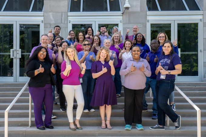 Arkansas department of education staff all dressed in purple, standing in front of the building, holding their thumbs up.