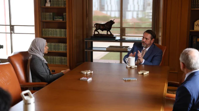 The image shows  President Jim Davis sitting across from a young woman at a table, speaking with his hands moving in front of him. A coffee mug and Texas-shaped coasters can be seen on the table with a bookshelf behind him. Photo by Justin Doud.