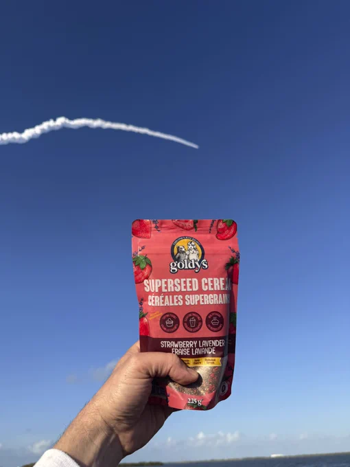 Hand holding a red Goldy's Superseed Cereal pouch with strawberry lavender flavor against a clear blue sky with a white vapor trail.