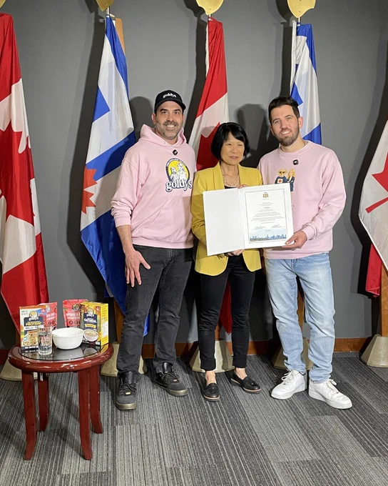 Toronto Mayor Olivia Chow and two individuals stand in front of Canadian and city of Toronto flags, holding an official certificate in a formal setting.