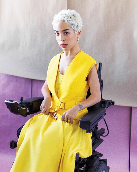 Professional portrait of Jilian Mercado, a woman with blonde hair in a vibrant yellow dress, seated in a black power wheelchair against a purple and beige background.
