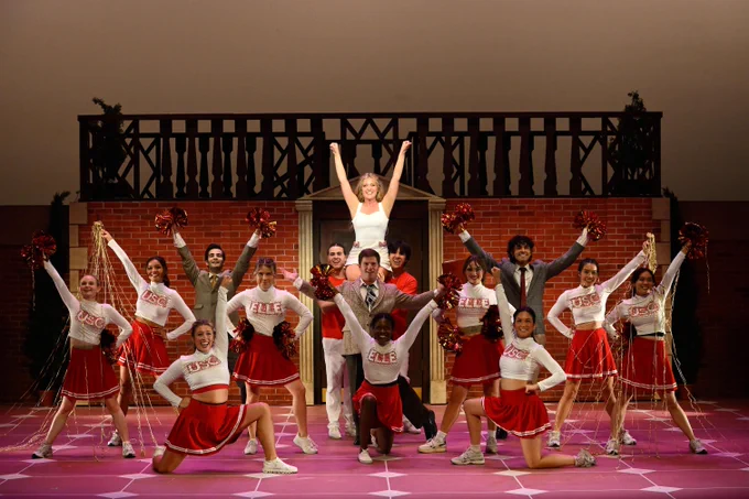 A group of cheerleaders and some men, presumably college students, are on stage with red pom poms. The cheerleaders are wearing red and white uniforms with \"USC\" or \"ELLE\" printed on them.