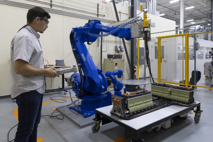 A person operates a large blue industrial robot arm in a factory setting, using a control device. Nearby, a table holds two battery modules. The environment includes machinery and safety equipment.