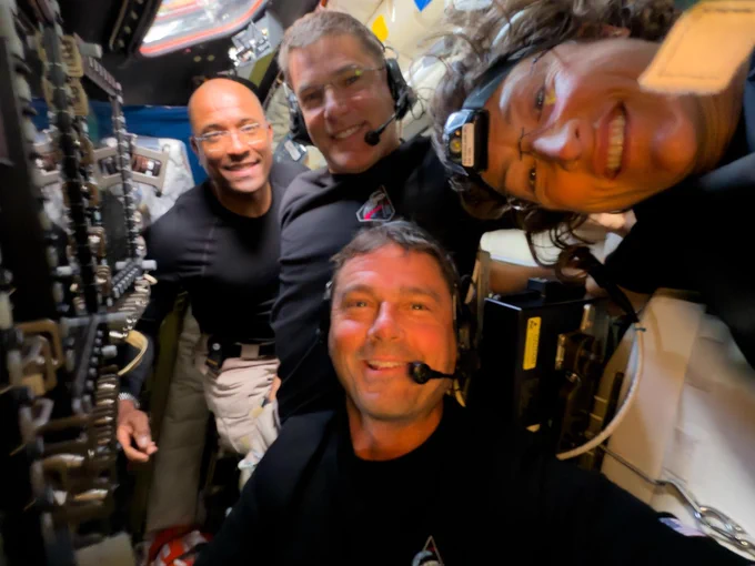 Four NASA astronauts are inside a spacecraft, smiling and wearing headsets. They are surrounded by various panels and equipment.