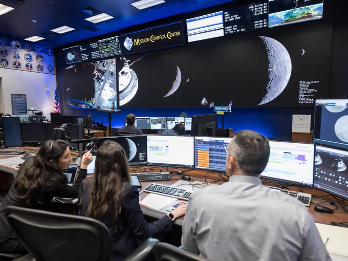 In a NASA control room, several people are seated at desks with multiple computer monitors. Large screens on the wall display images of a spacecraft, the Moon, and space-related data. The room is identified as the Mission Control Center.