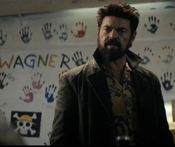 Billy Butcher wearing his trench coat and Hawaiian shirt, looking at Starlight. Behind him is a white board with text that reads Wagner with kids handprints and a skull and crossbones
