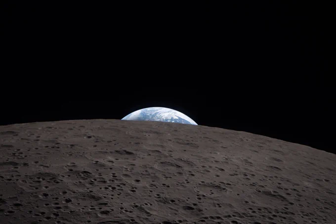 Earth peeks just above the horizon of the Moon, which looms large in the foreground. The Moon's surface is covered in craters of various sizes, giving it a spongy, bumpy texture. Earth's upper edge is a dreamy light blue with white clouds.