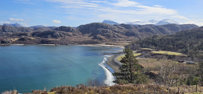 A pebble and yellow sand beach with turquoise water, and rounded mountains in the background.