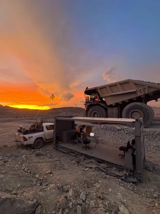 A large mining truck on a gravel road at sunset, casting a silhouette against a vibrant sky. In the foreground, a small open structure with desks and a laptop is set up beside a pickup truck.