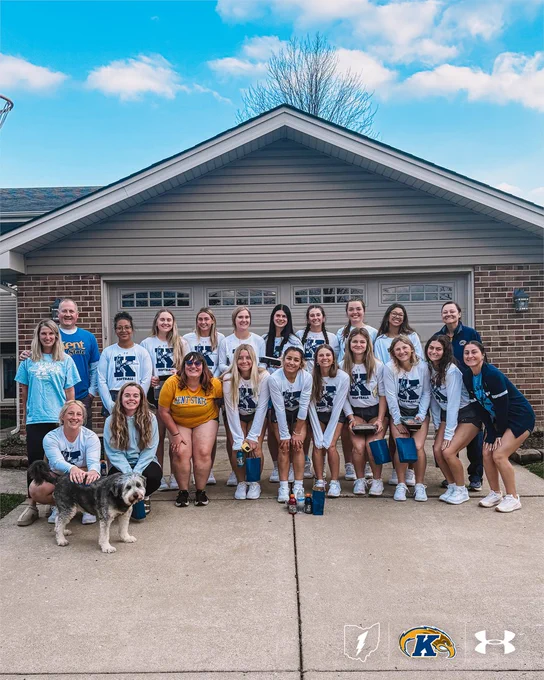 Players and coaches on the Kent State softball team are wearing Kent State-themed attire, standing and kneeling in front of a house. A dog is sitting at the front left. Logos of Kent State and Under Armour are visible at the bottom of the image.