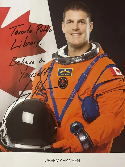 Astronaut Jeremy Hansen in an orange space suit holding a helmet, standing before a Canadian flag backdrop. Singed with the words Toronto Public Library Believe in Yourself