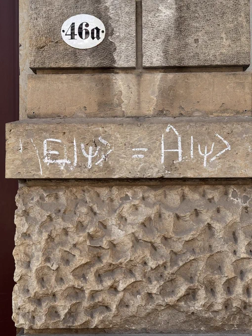 Close-up of a sandstone building facade showing the equation “Ĥ|ψ⟩ = E|ψ⟩” written in white chalk on a horizontal stone ledge. The stone below has a rough, pitted texture. Above it, an oval enamel house number plate reads “46a” in black Fraktur script. A dark red door frame is visible on the left edge.