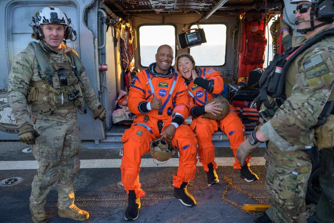 Artemis II astronauts Victor Glover and Christina Koch, wearing orange flight suits, sit on the edge of a helicopter, flanked by uniformed personnel. Both astronauts are grinning. Credit: NASA/Bill Ingalls