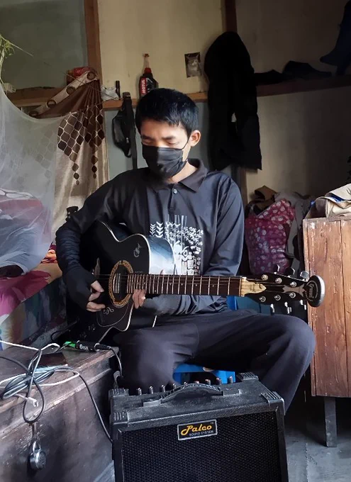 "A young man from Aibawk, Mizoram, wearing a black mask and playing an acoustic-electric guitar through an amplifier. He is focused on a guitar solo. Photo by Chhanchhana Zote Hmar."