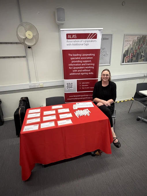 A woman dressed in black is sat at a table there is a table to her right with a red tablecloth on top, covered with information cards and goodies