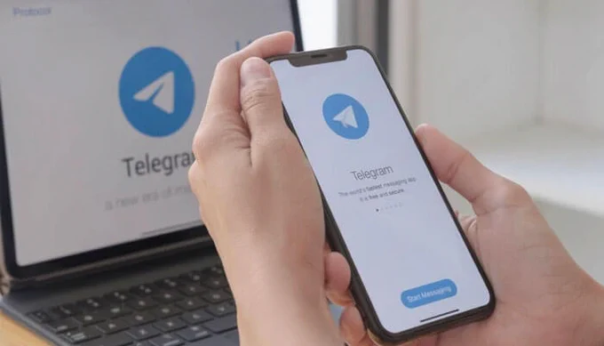 Person holding an iPhone with the Telegram app open, displaying the app’s welcome screen with the 'Start Messaging' button and Telegram logo. A laptop with the Telegram logo is visible in the background.