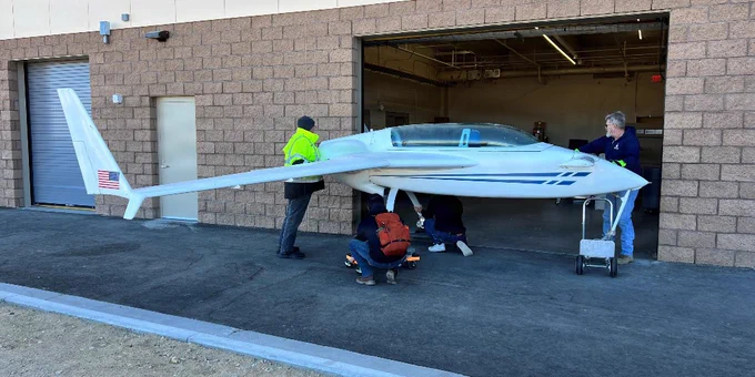 A sleek white glider is being inspected by people outside a hangar with a brick facade.