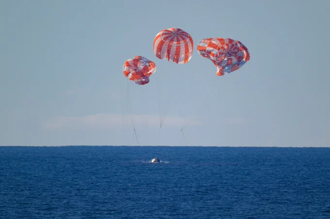 Three red and white striped parachutes deployed above a spacecraft landing in the ocean during daylight.