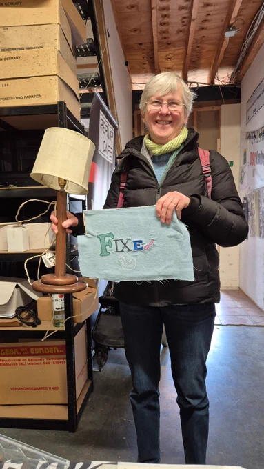 A person in a workshop holds a wooden lamp in one hand and a blue embroidered cloth reading “FIXEd” in the other. Shelves of boxes and posters fill the background