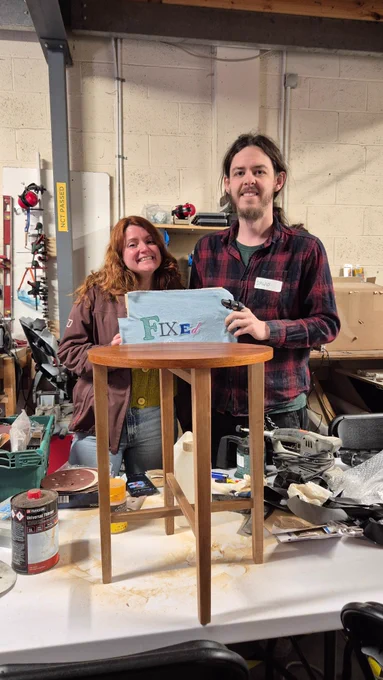 Two people stand in a workshop behind a newly repaired wooden stool. One holds a power tool while both display a colourful “FIXEd” cloth. Tools and materials line the shelves behind them.