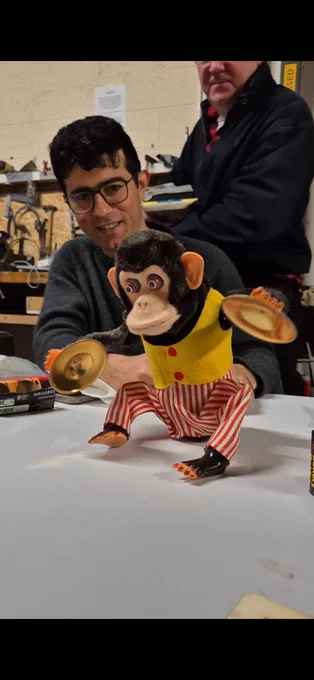 A vintage cymbal‑banging toy monkey sits on a workbench in a busy workshop. Two people stand behind it, partially visible, surrounded by tools and equipment.