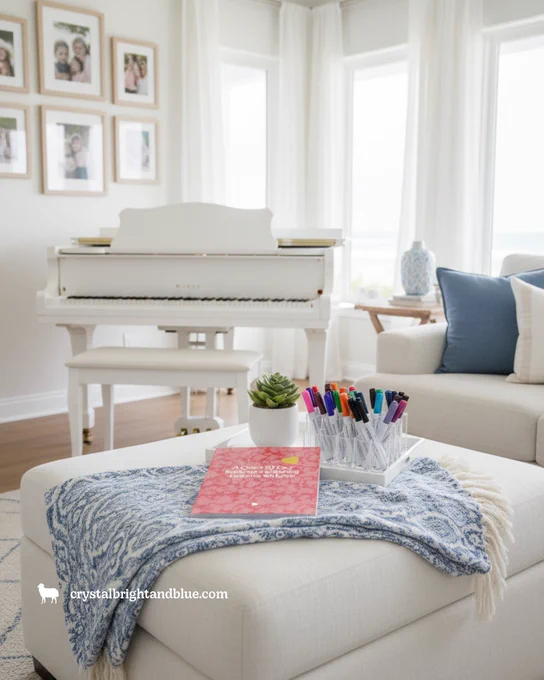 Coral pink "A Quiet 30-Day Scripture Coloring Journey on Love" book on white linen ottoman with blue paisley throw blanket, clear cup of colorful markers, white succulent pot, white grand piano, blue accent pillows.