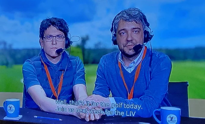 Two men, acting as sports commentators, are sitting at a desk and holding hands. The man on the left has dark hair and glasses and is wearing a blue vest over a blue shirt. The man on the right has salt-and-pepper hair and is wearing a blue sweater over a light-colored collared shirt. Both men are wearing headsets with microphones and orange lanyards. In front of them are blue mugs and a pencil. The background is a green field under a blue sky. Subtitles at the bottom read, "Well, that's it for the golf today. Join us next week for the LIV".
