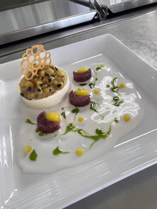 A square white plate featuring a decorative dessert. On the left is a round serving with diced fruit and a lattice garnish. The right side has artistic dollops of yellow sauce and small pieces of fruit arranged on a creamy white base with green accents.
